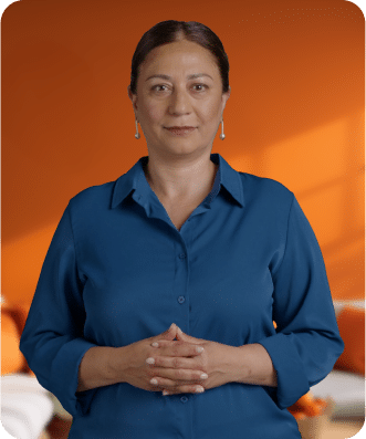 Woman with brown hair pulled back, wearing a blue button-up shirt, stands indoors against an orange background with hands clasped.