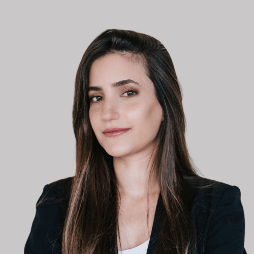A woman with long brown hair wearing a black blazer and white top poses against a plain light grey background. Noa Sella